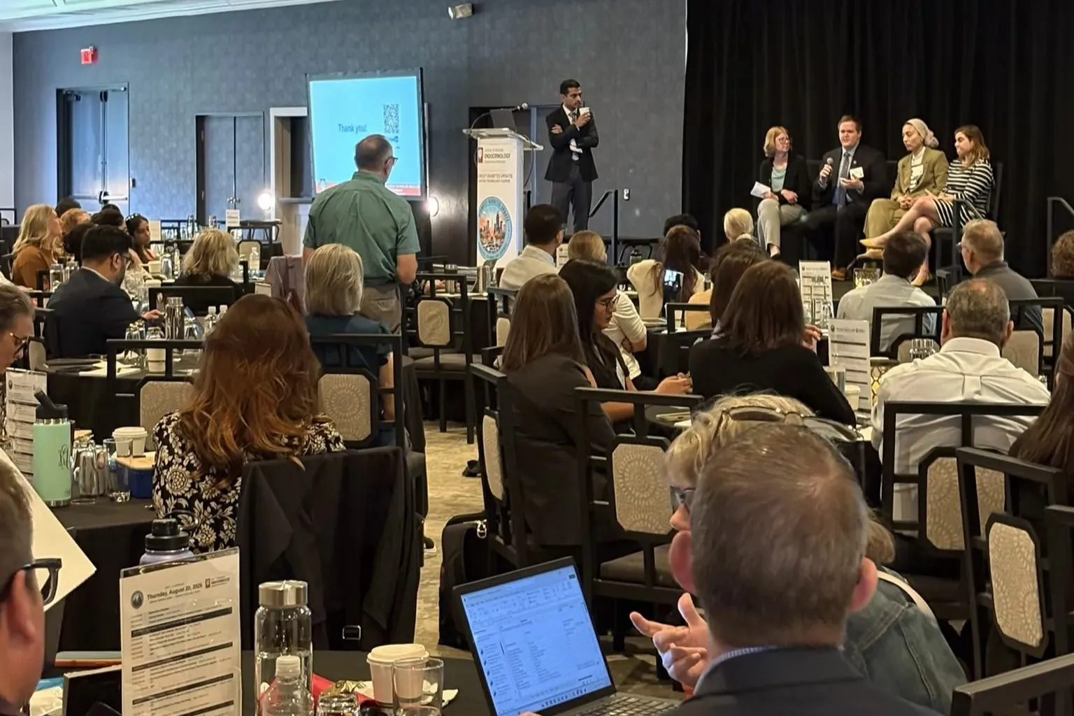 A group of people sit in a conference room watching a person ask a question of a panel of people during a presentation.