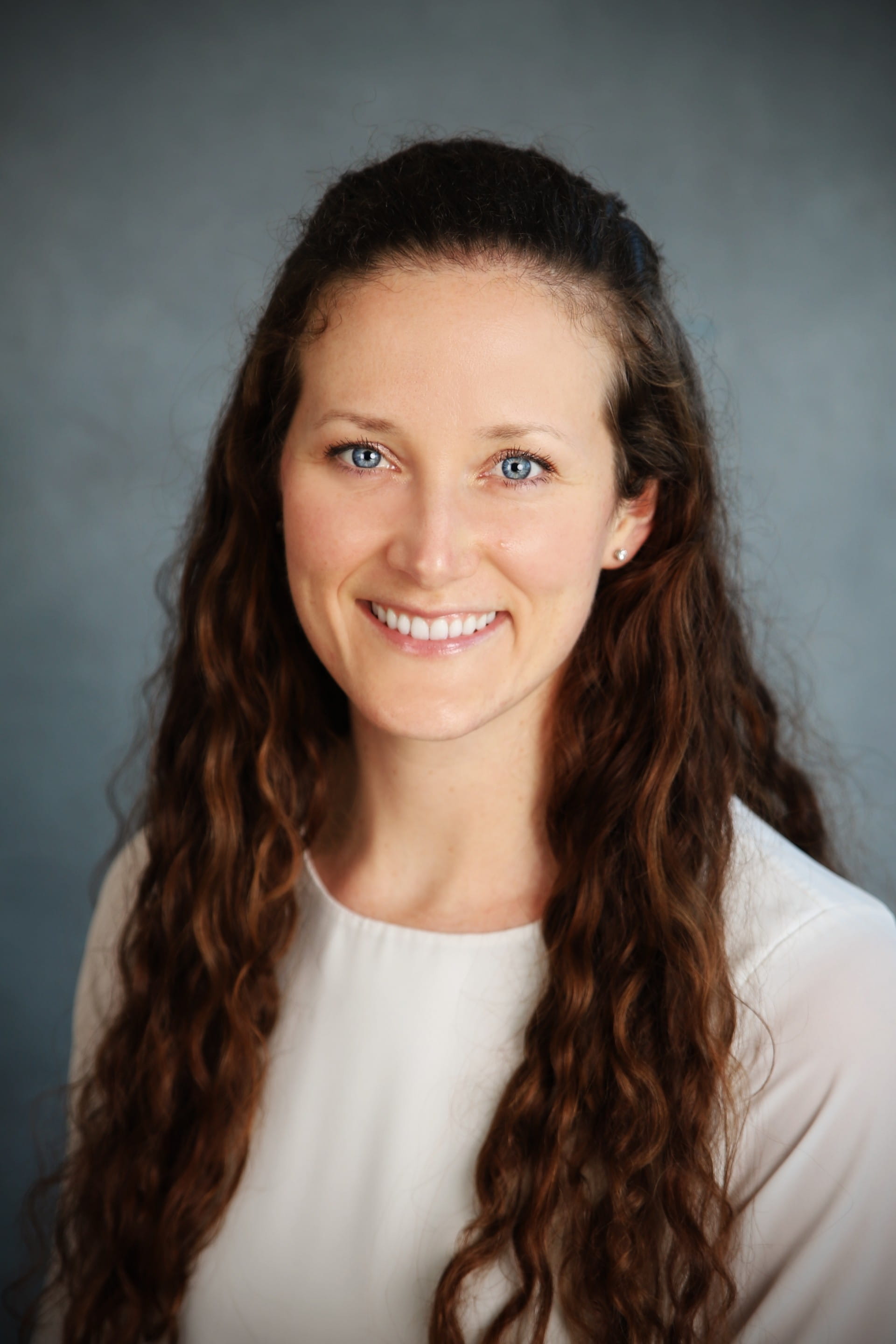 Woman with long, brown, curly hair is wearing a white shirt and smiling.
