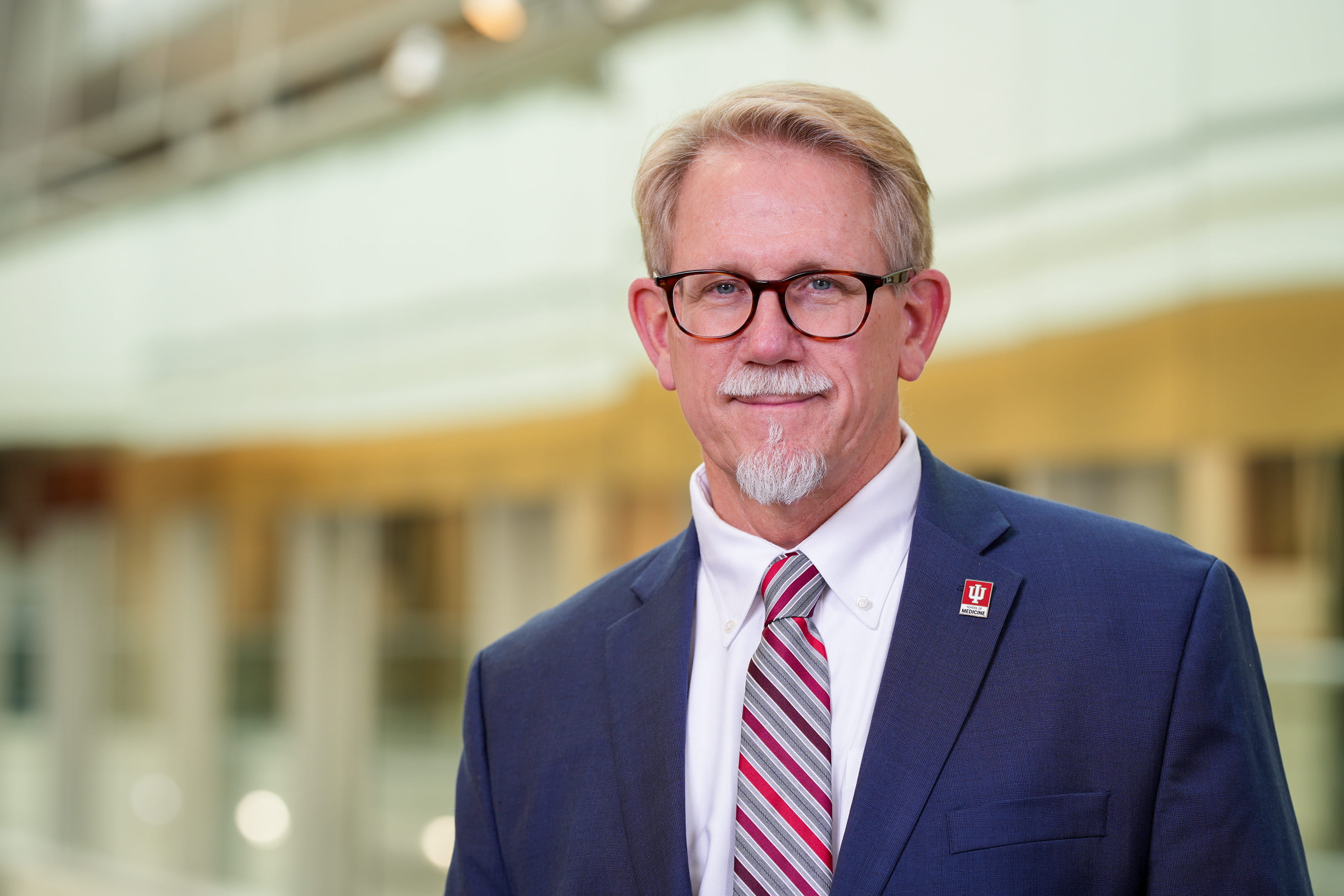 portrait of brad allen in a building lobby. he's wearing a dark suit, red striped tie and glasses