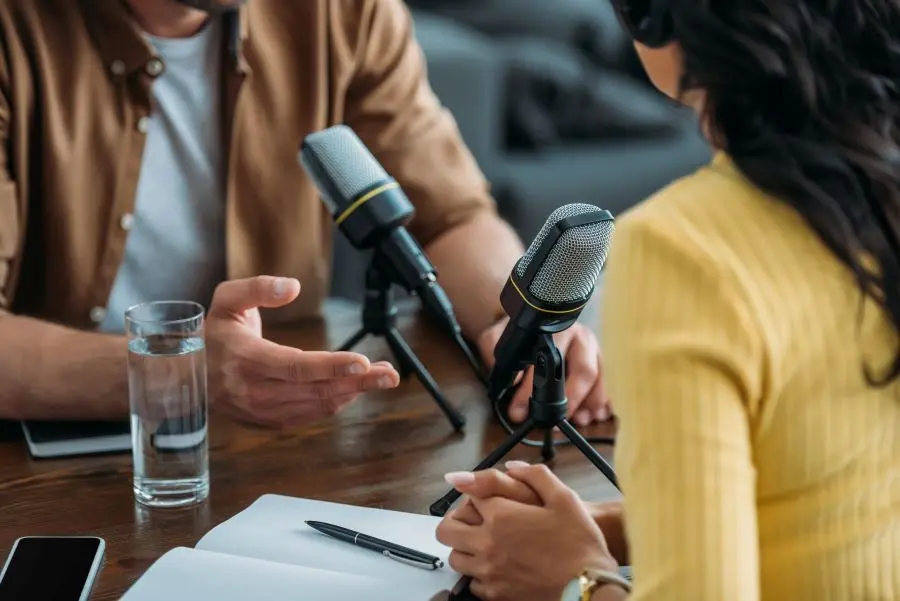 two people sit at a table in front of microphones to record a podcast