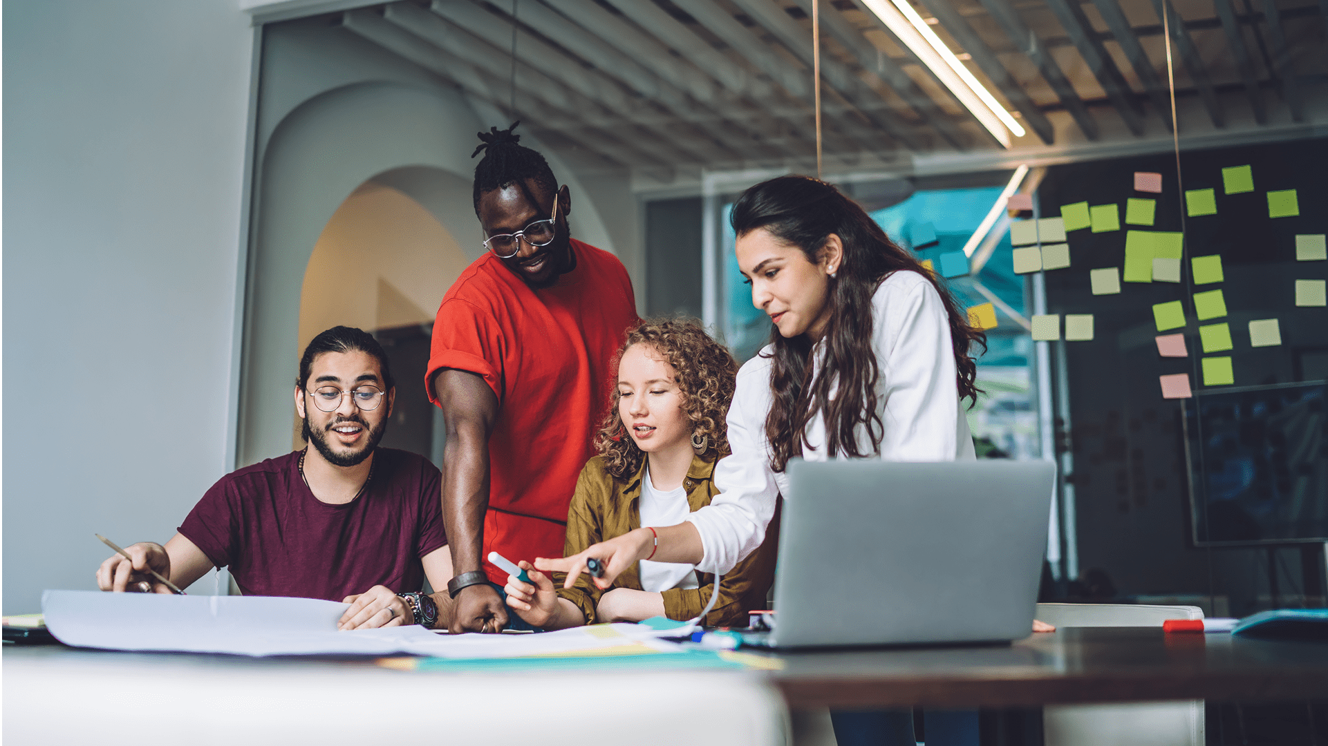 A diverse group of four professionals collaborating around a table with documents and a laptop in a modern office featuring glass walls and sticky notes.