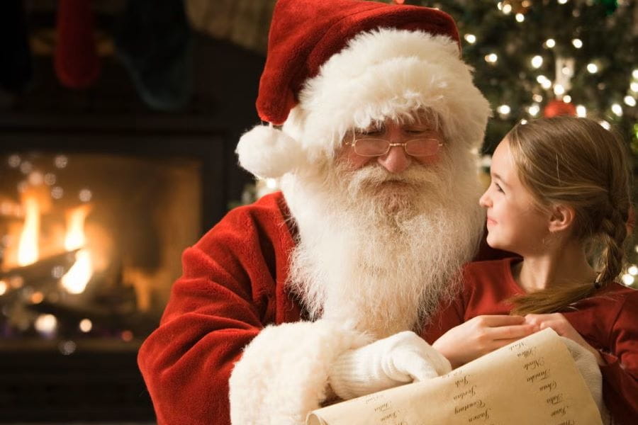 a girl siting in Santa's lap, smiling at him, with Christmas lights and fireplace in the background
