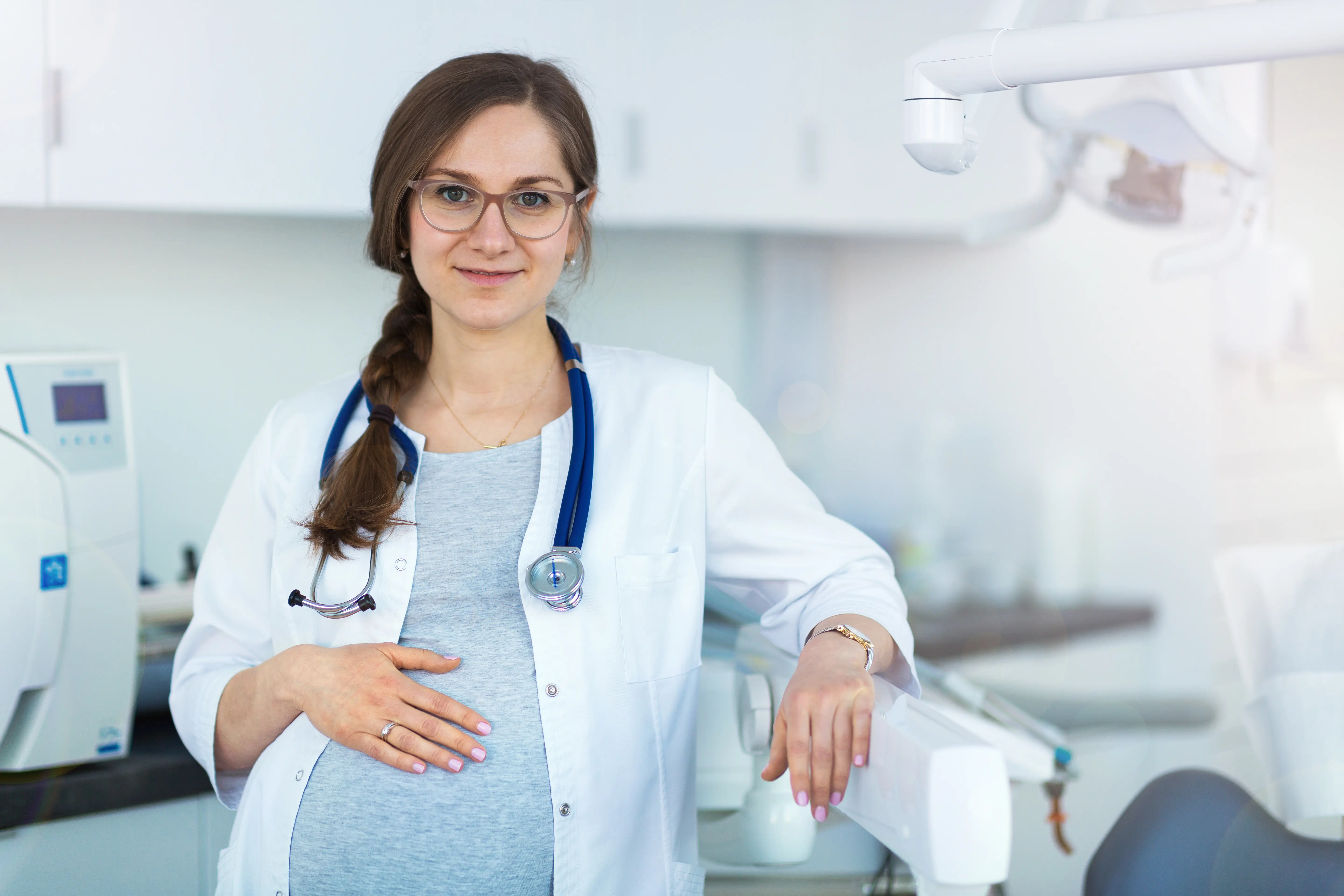 A smiling, pregnant physician in a white coat and stethoscope looks toward the camera with her hand resting on her belly.