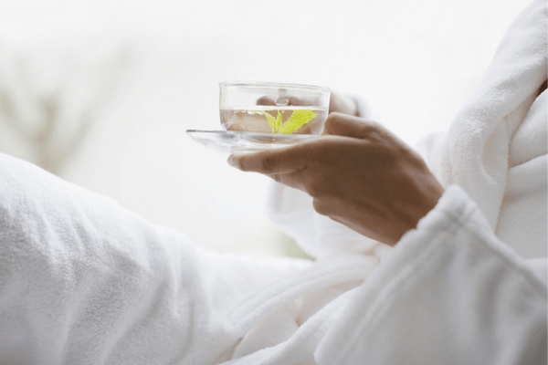 Person in white robe holding a clear glass with tea and a green leaf