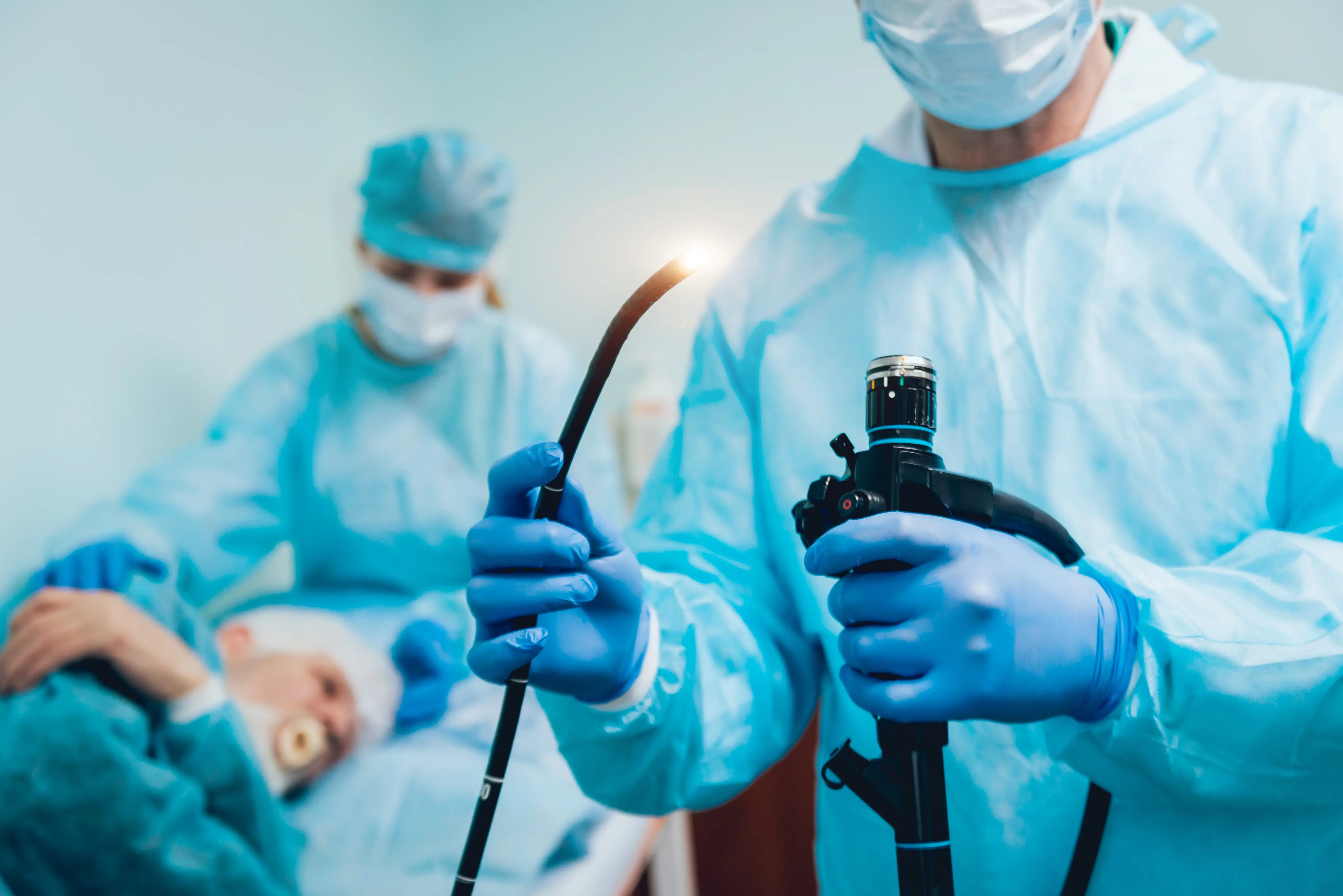 Foreground: A doctor in scrubs holds an endoscope with a light. Background: A clinician preps a patient for an endoscopic procedure.