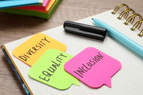 Notebook open on a wood table, with three speech bubble-shaped post-it notes with one word on each- "Diversity" on orange, "Equality" on green and "Inclusion" on pink. A blue pen with a black cap laying next to it is on the notebook next to the notes