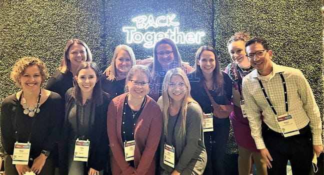 A group of ten colleagues pose together in two rows in front of a hedge backdrop. The neon sign behind them says "BACK TOGETHER." They are all smiling and wearing lanyards with nametags.
