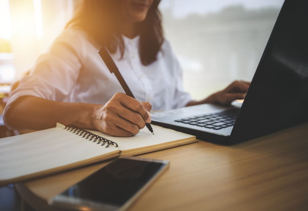 Woman sits at a laptop and writes in a notebook