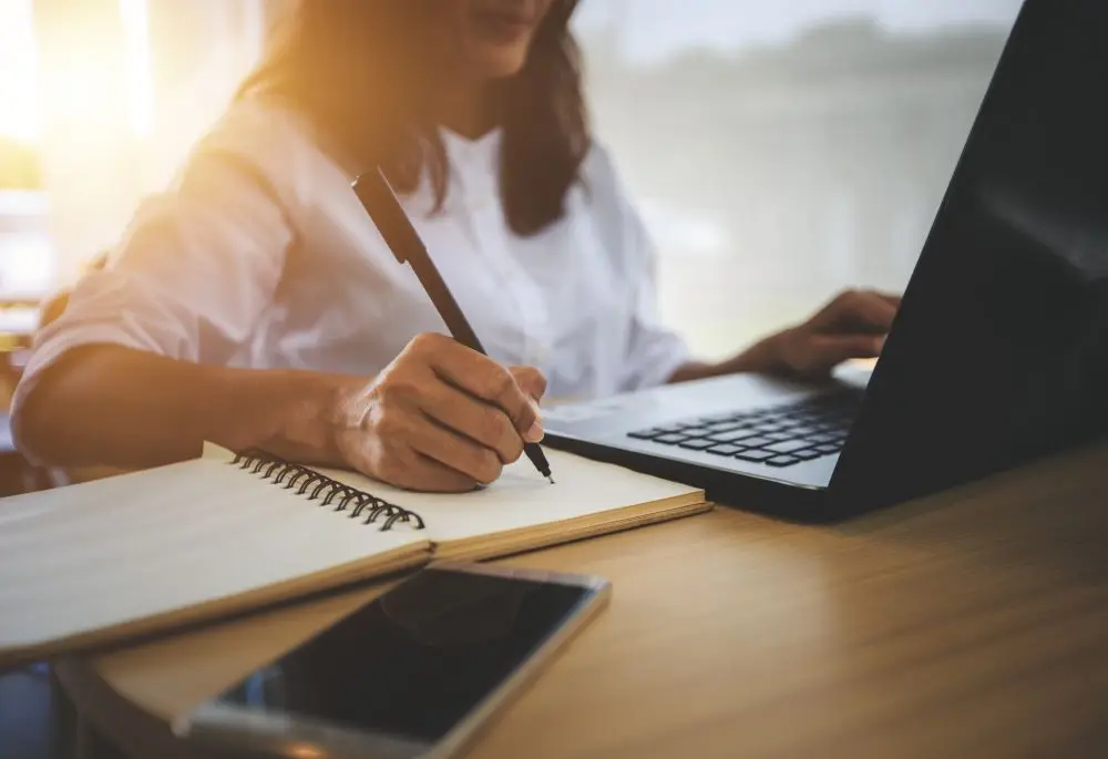 Woman sits at a laptop and writes in a notebook