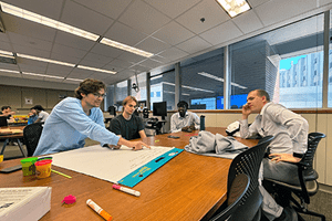 Three AIM SIG students sit around a table discussing a project on a board.