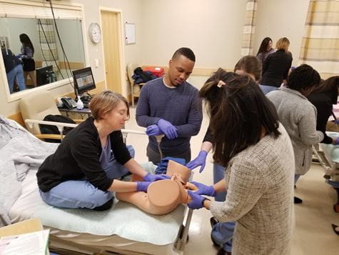 OB-GYN trainees work with a manikin during training Three trainees work on a manikin while a female faculty member supervises, sitting on hospital bed in simulation center