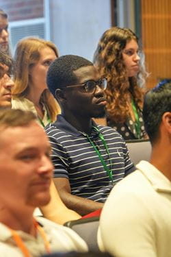 Mout-Maine Moustapha, Black man wearing glasses and striped polo, sits among peers and listens during an orientation presentation for new medical students.