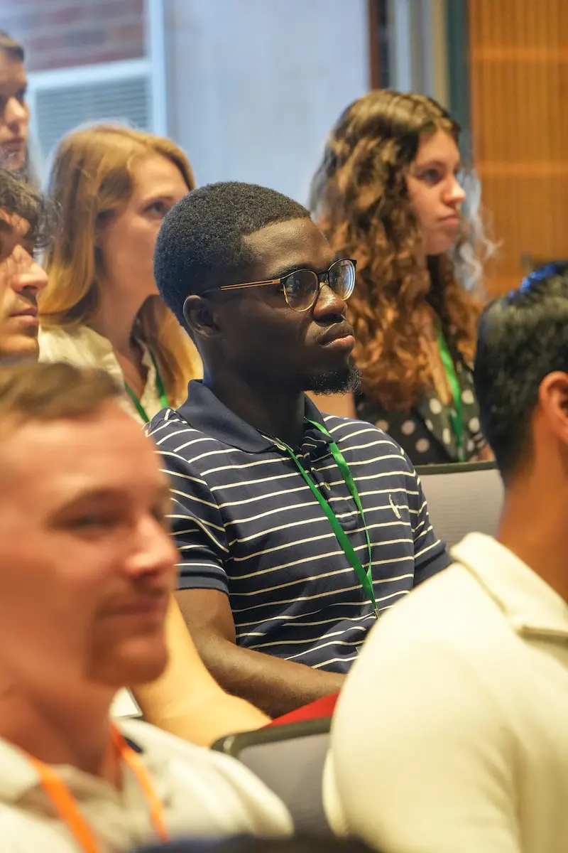 Mout-Maine Moustapha, Black man wearing glasses and striped polo, sits among peers and listens during an orientation presentation for new medical students.