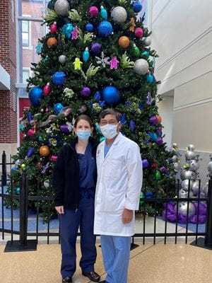 Michele and Chan Saysana in their scrubs standing in front of a tall Christmas tree in the Riley Hospital lobby