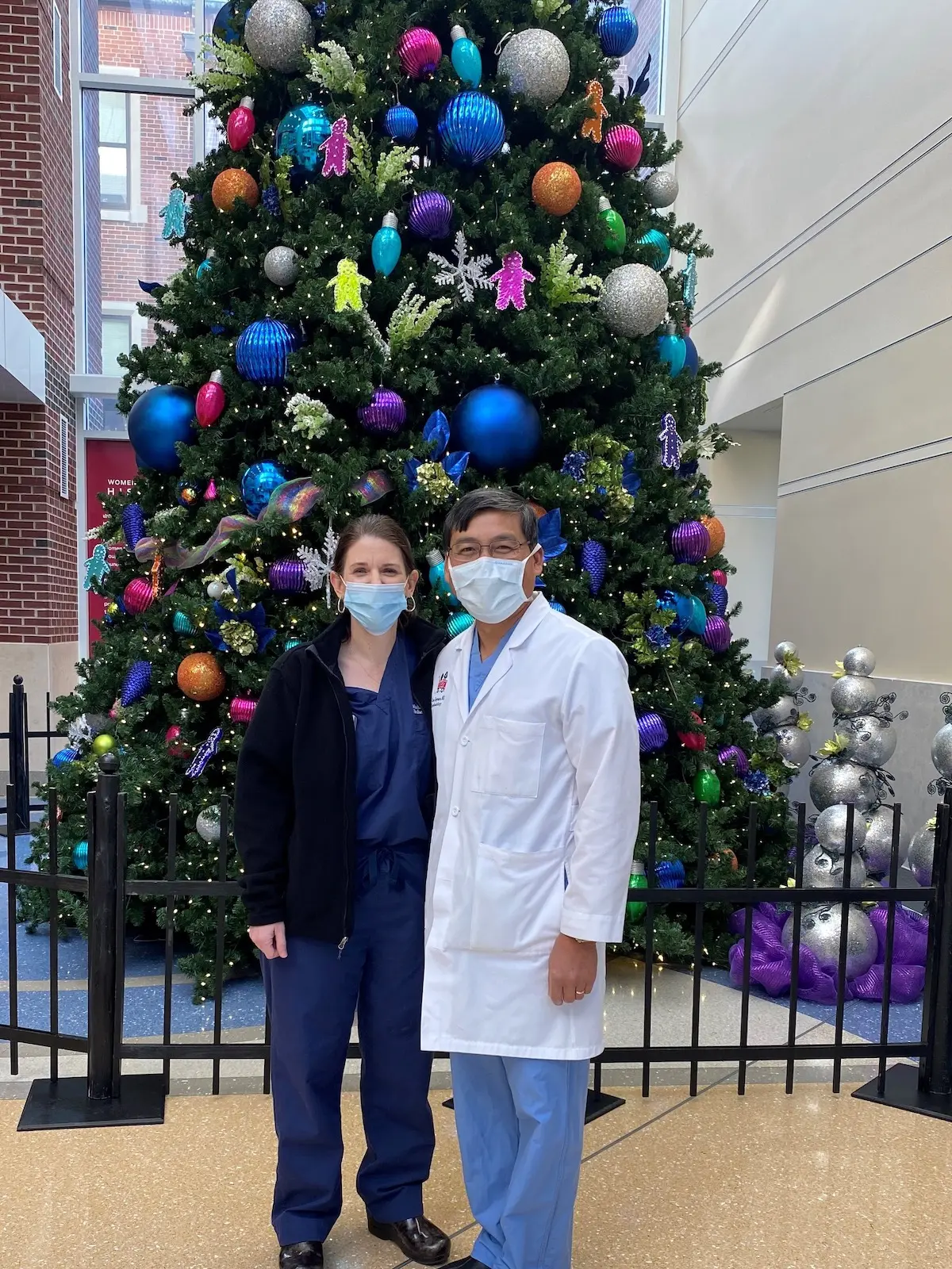 Michele and Chan Saysana in their scrubs standing in front of a tall Christmas tree in the Riley Hospital lobby