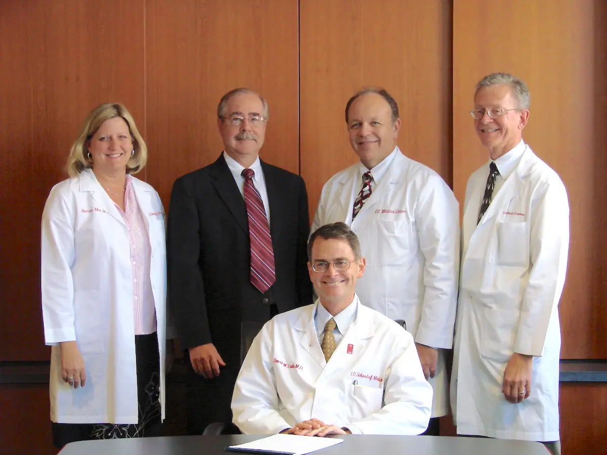 Dr David Crabb seated at table with Department of Medicine leaders standing behind him, including Charlie Fox in black suit, circa 2010s