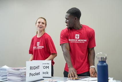 Mout-Maine Moustapha, young Black man wearing red IU School of Medicine T-shirt, with a blonde female classmate in matching shirt, sorting piles of paperwork at a table during community service event