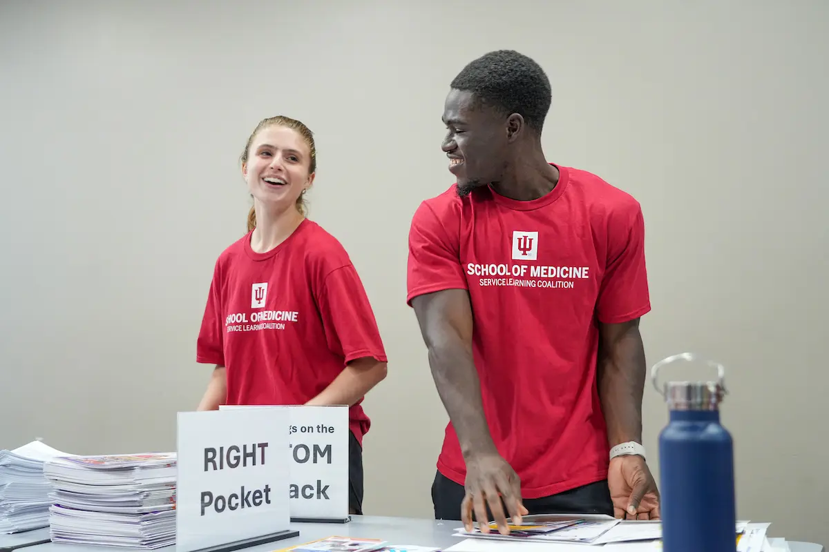 Mout-Maine Moustapha, young Black man wearing red IU School of Medicine T-shirt, with a blonde female classmate in matching shirt, sorting piles of paperwork at a table during community service event
