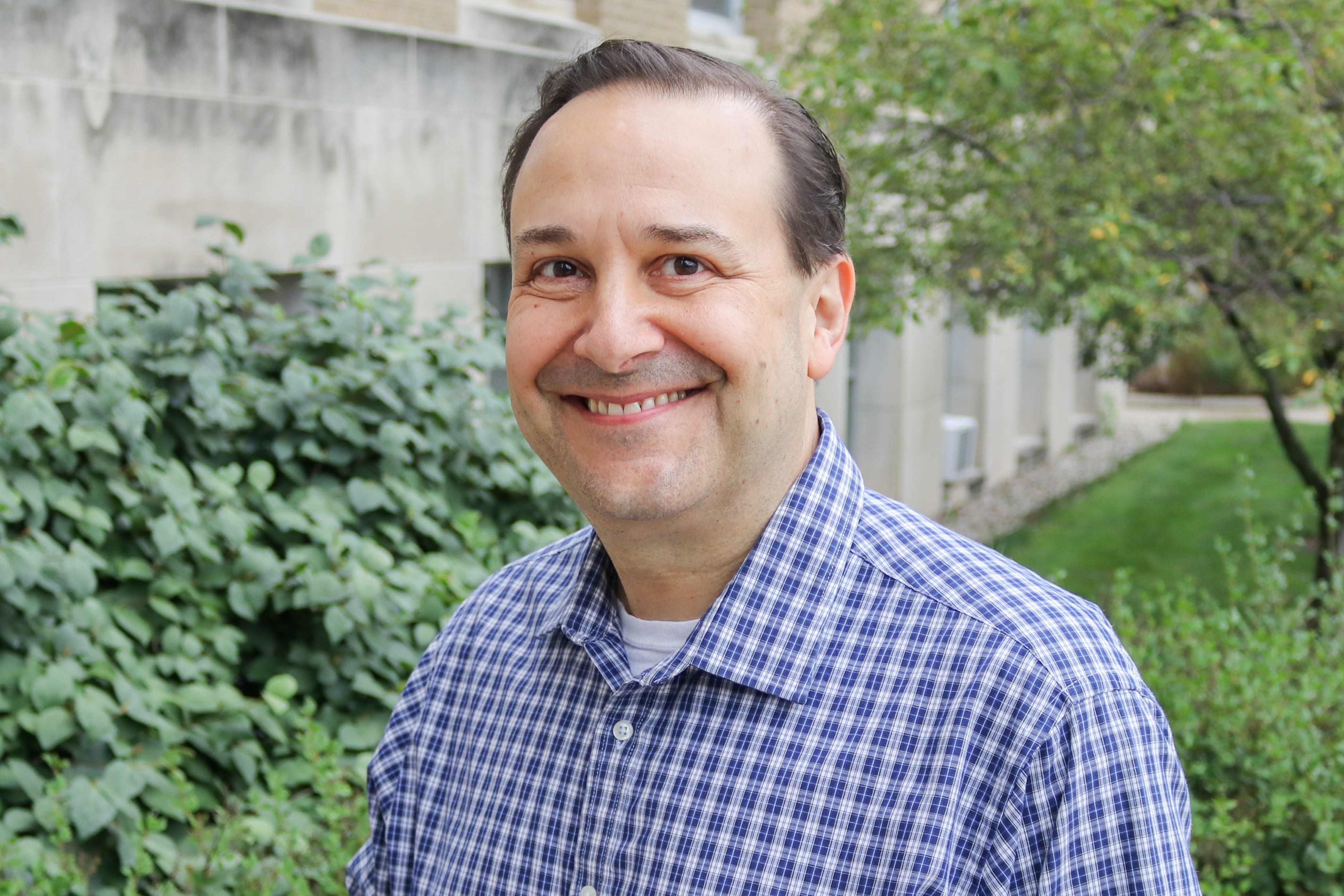David Haas, MD, smiles for his headshot outside. Behind him, there are trees and bushes obscuring a limestone building.