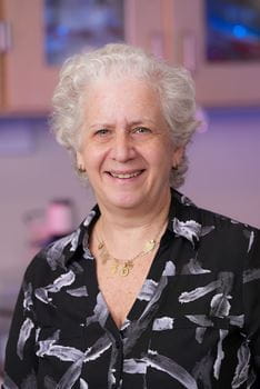 Lilian Plotkin, woman with short white hair wearing black printed blouse in laboratory with purple lighting