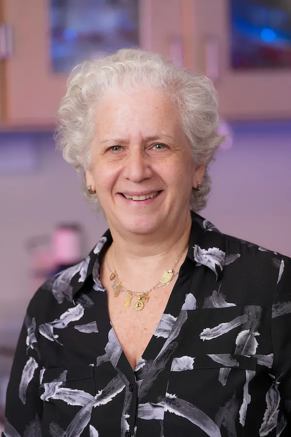 Lilian Plotkin, woman with short white hair wearing black printed blouse in laboratory with purple lighting