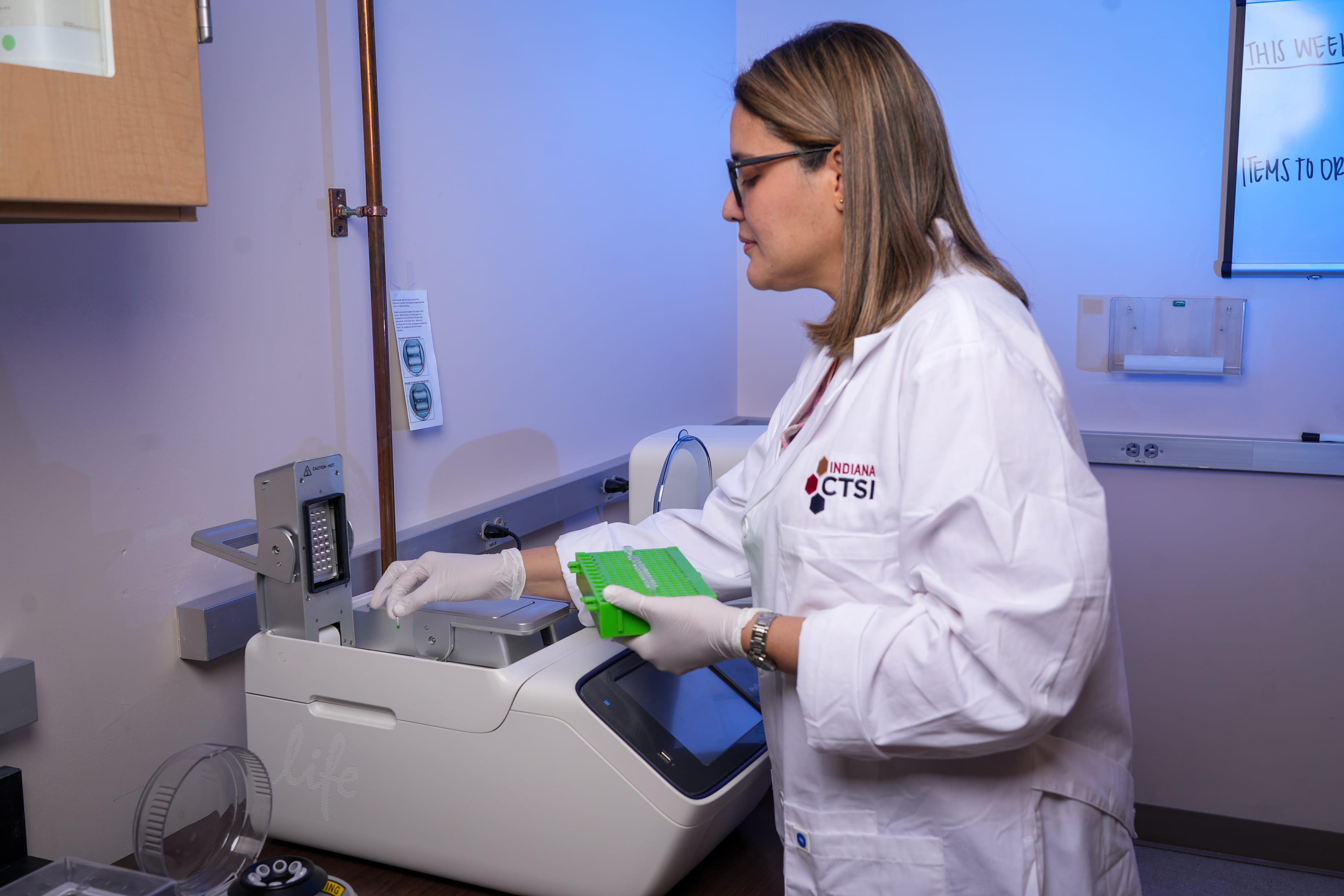 Roquelina Pianeta, woman in white coat, works with a machine in the Plotkin Lab.