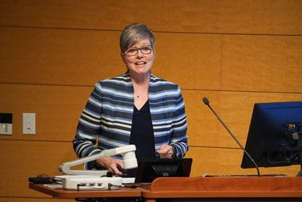 Mary Dankoski stands at wooden desk with microphone, speaking during the Fall 2024 All School Meeting