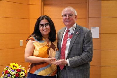 Leila Ekbia, in colorful blouse and glasses, holding award and posing with Dean Hess