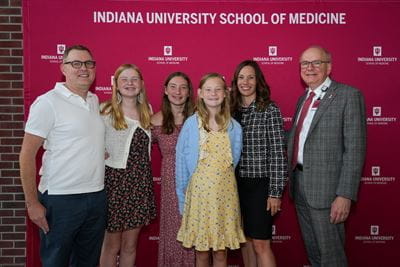 Cynthia Holladay poses with her family group and Dean Jay Hess in front of a red, IU School of Medicine backdrop