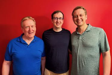 Richard Gunderman, white man in blue polo shirt; Peter Gunderman, man with glasses in navy shirt; and David Gunderman, tall man in polo shirt, pose together in front of red wall