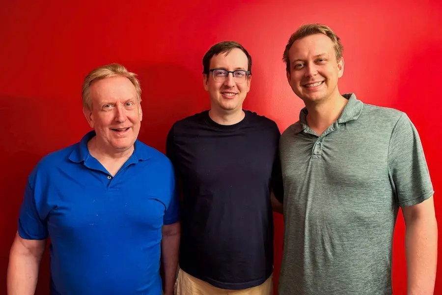 Richard Gunderman, white man in blue polo shirt; Peter Gunderman, man with glasses in navy shirt; and David Gunderman, tall man in polo shirt, pose together in front of red wall