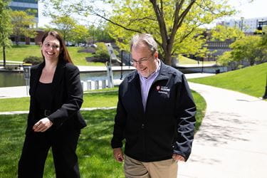 Andrea Ligler and Michael Feldman, both in business attire, walk along an outdoor path on a sunny day