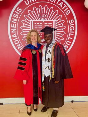 Mout-Maine Moustapha poses with IU President Pamela Whitten in front of red wall with the IU seal, both wearing graduation regalia