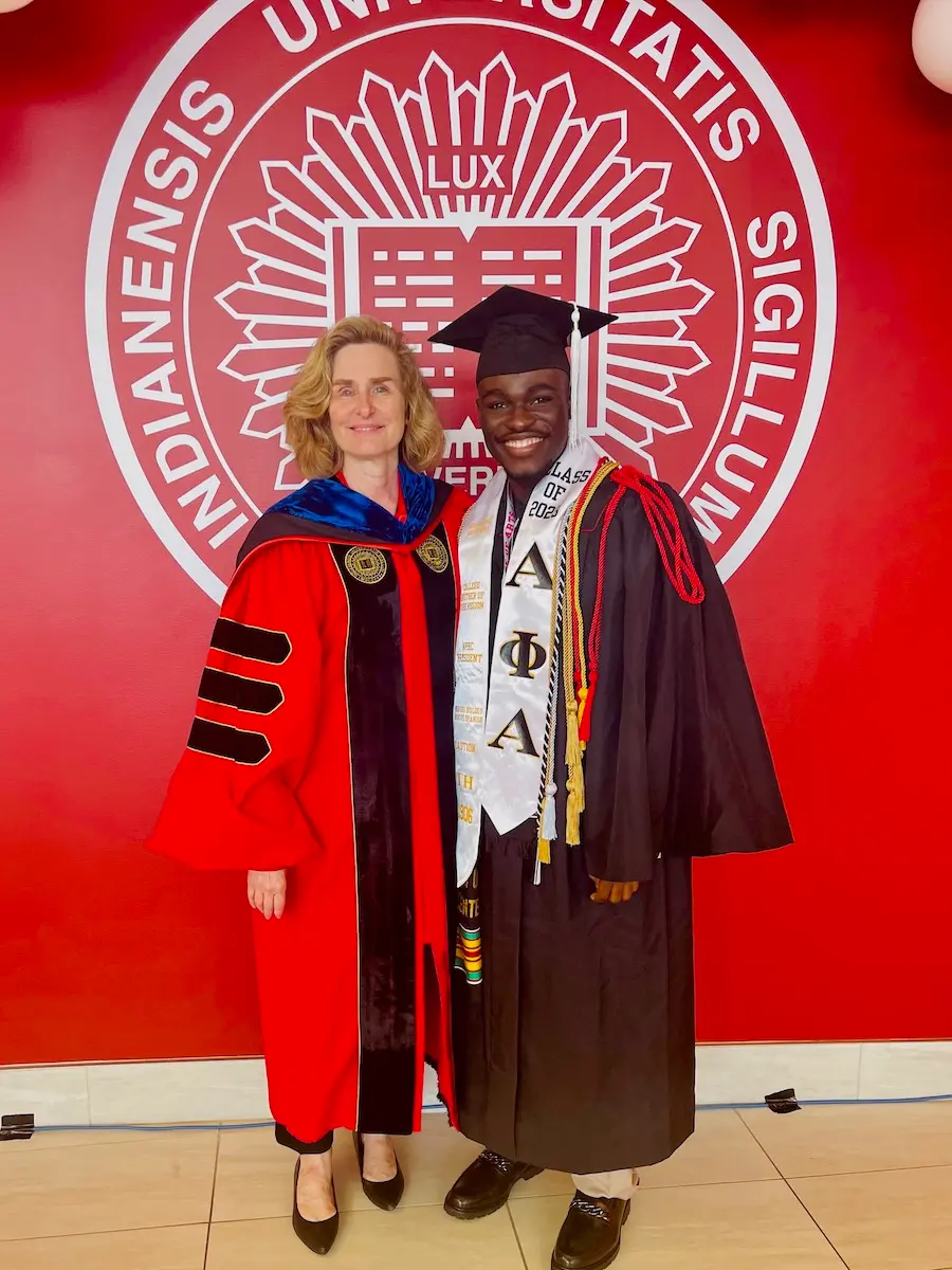 Mout-Maine Moustapha poses with IU President Pamela Whitten in front of red wall with the IU seal, both wearing graduation regalia