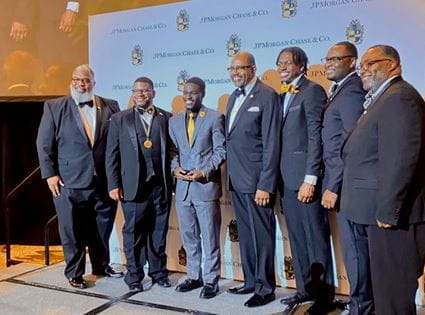 Mout-Maine Moustapha, in gray suit and yellow tie, holding trophy award, standing with six Black men in dark suits on stage