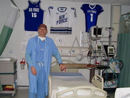 Dr. Ritter wearing surgical scrubs standing beside a hospital bed with US Air Force jerseys hanging on the wall