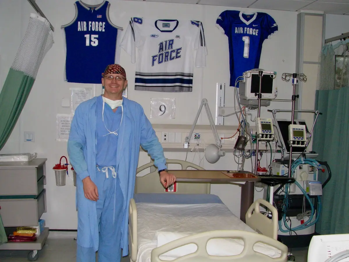 Dr. Ritter wearing surgical scrubs standing beside a hospital bed with US Air Force jerseys hanging on the wall