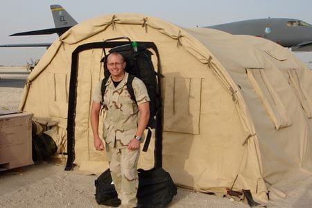 Dr. Ritter in military fatigues wearing a large backpack and standing in front of a military tent and airplane