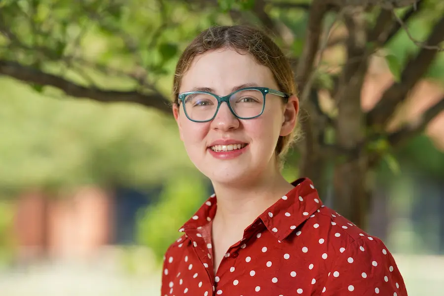 Sarah Vaught, white female, blue glasses, wavy hair in pony tail, red polka dot button-up shirt, outside under a tree