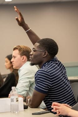 Mout-Maine Moustapha, Black man in striped shirt, raises his hand to speak during an orientation session with incoming medical students.