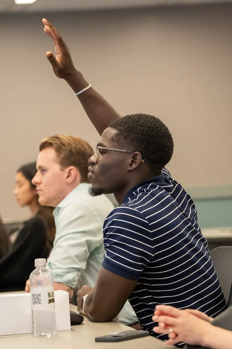 Mout-Maine Moustapha, Black man in striped shirt, raises his hand to speak during an orientation session with incoming medical students.
