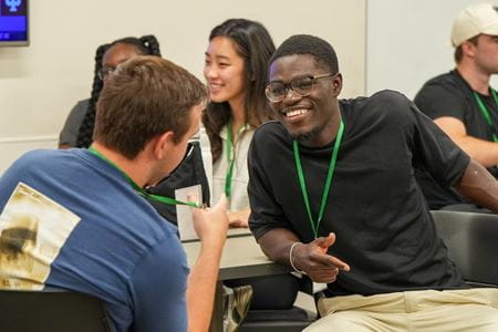 Mout-Maine Moustapha, Black man wearing glasses and a dark shirt, smiling while talking with a male classmate during orientation.