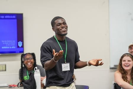 Mout-Maine Moustapha, Black man wearing black sweater and name badge lanyard, gestures as he speaks in front of laughing classmates during an orientation activity