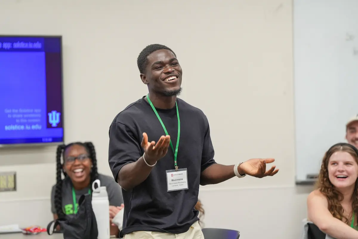 Mout-Maine Moustapha, Black man wearing black sweater and name badge lanyard, gestures as he speaks in front of laughing classmates during an orientation activity