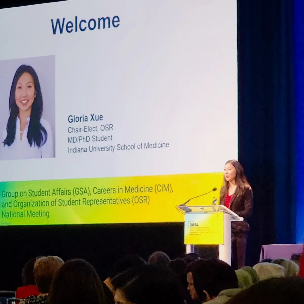 Gloria Xue speaks from a podium onstage with a slide of her headshot and bio in the background.