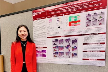 Gloria Xue, wearing red jacket, stands by a poster presentation of her research