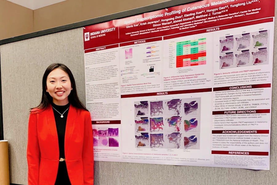 Gloria Xue, wearing red jacket, stands by a poster presentation of her research