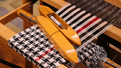 Weaving tools are laid out on top of a red, black, and white woven piece that is a work-in-progress.