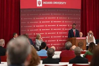 IU medical student Myke Spencer speaks at the Medical Education and Research Building dedication as IU President Pamela Whitten and others listen. Red curtain and red Indiana University logo background behind podium