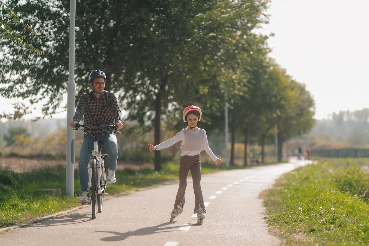 A dad on a bike next to a girl on rollerblades, traveling on a bike path